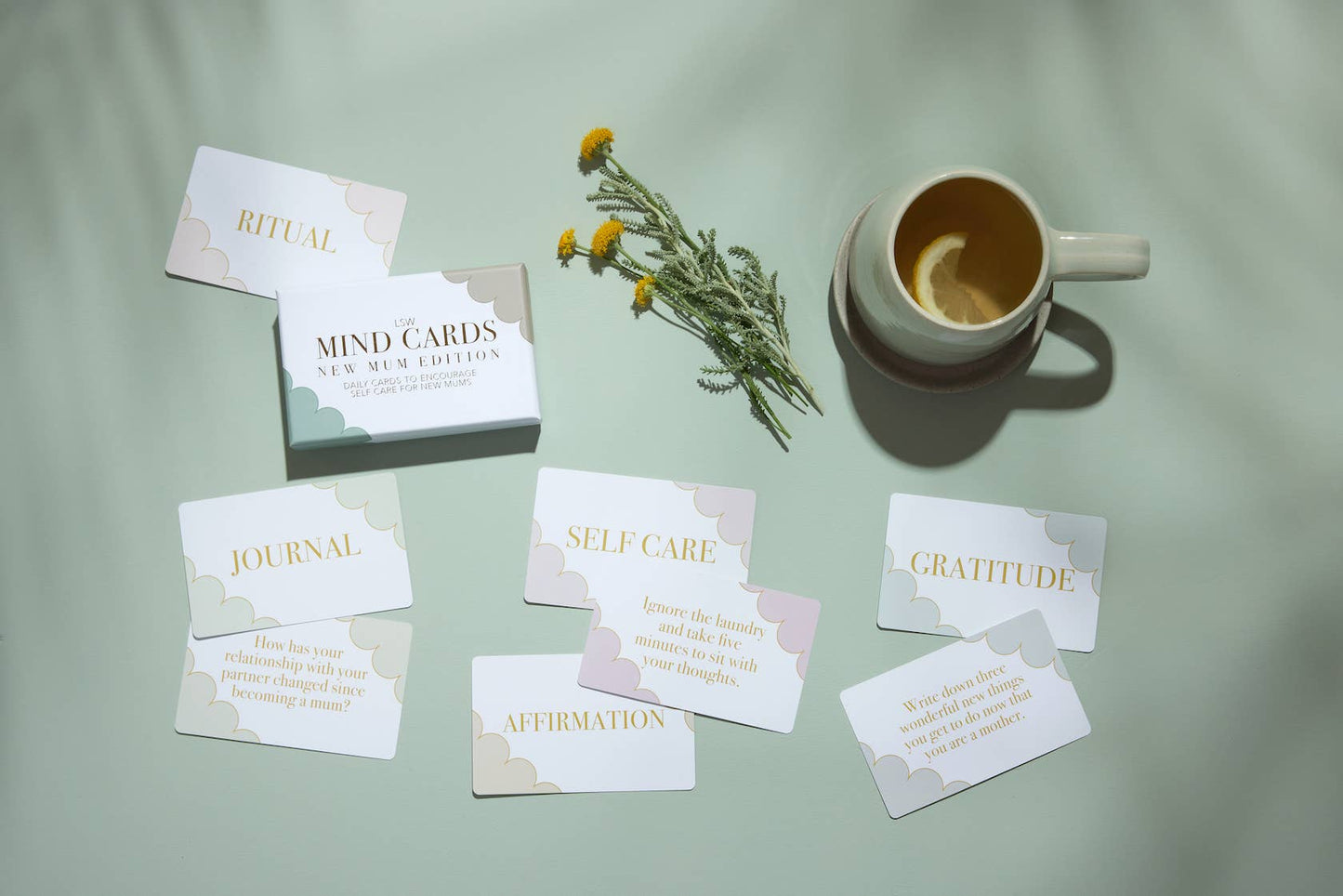 Set of self-care cards on a light green surface with a mug of tea and flowers.