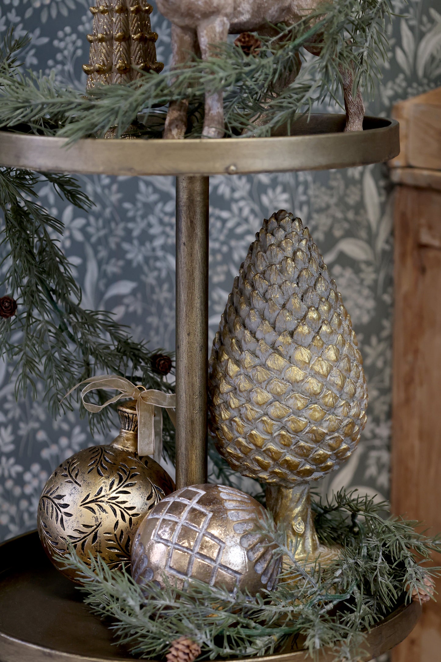 Decorative tiered stand with gold ornaments and greenery against a patterned wall.