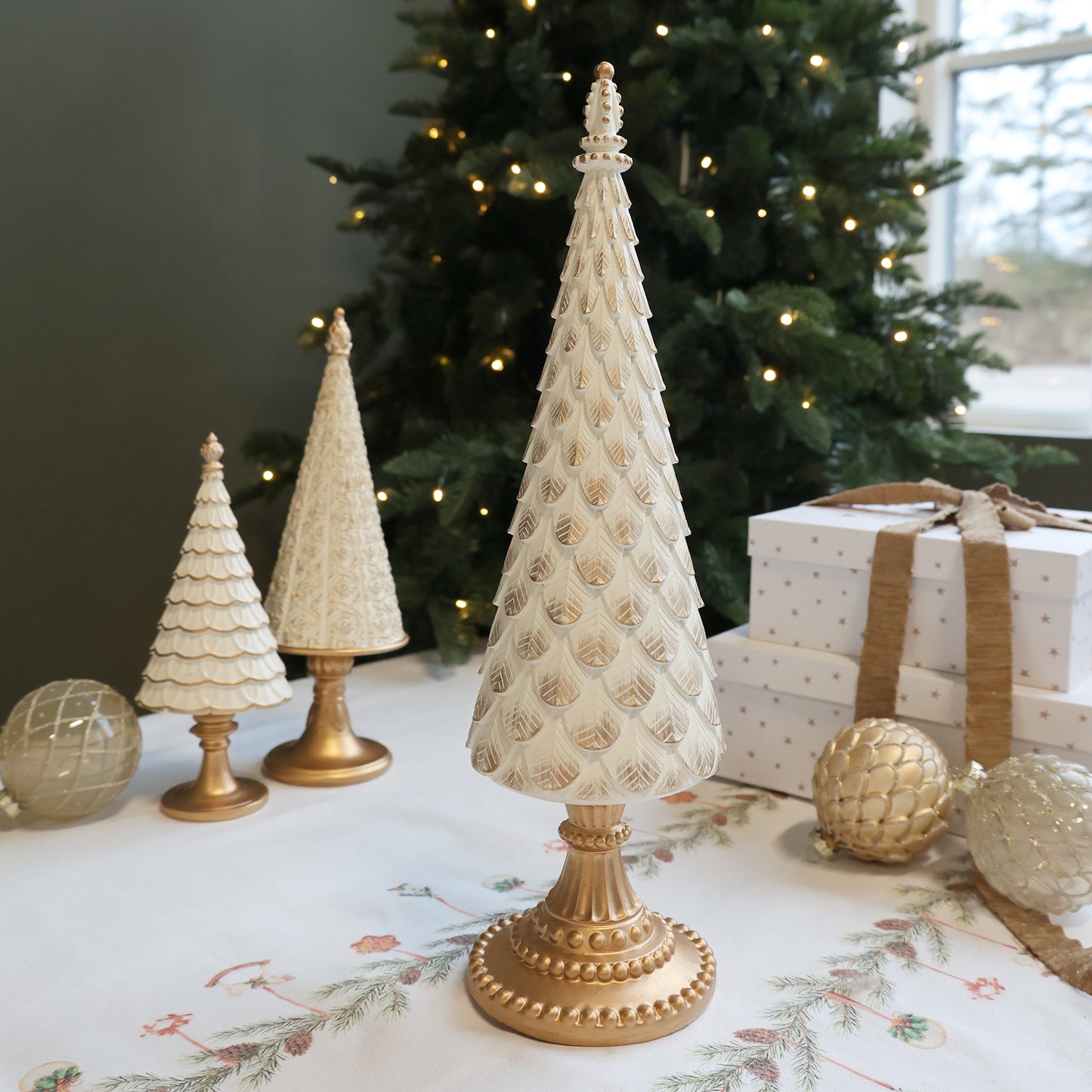 Decorative Christmas trees with gold bases on a table in front of a decorated tree.