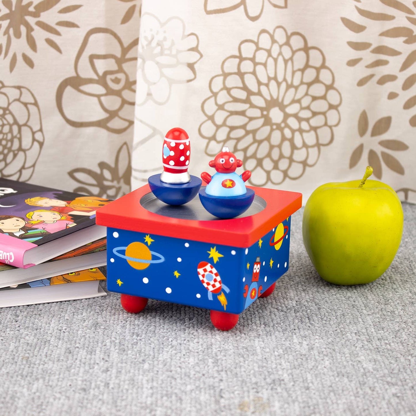 Colorful toy with characters on a stand next to an apple and books against a floral-patterned wall.