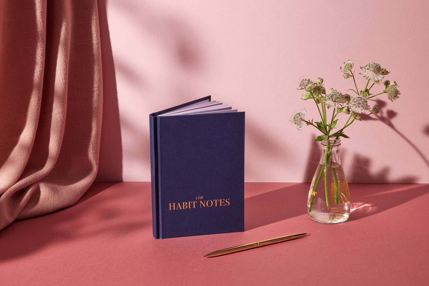 Blue notebook titled 'Habit Notes' with a pen and small vase of flowers on a pink surface.