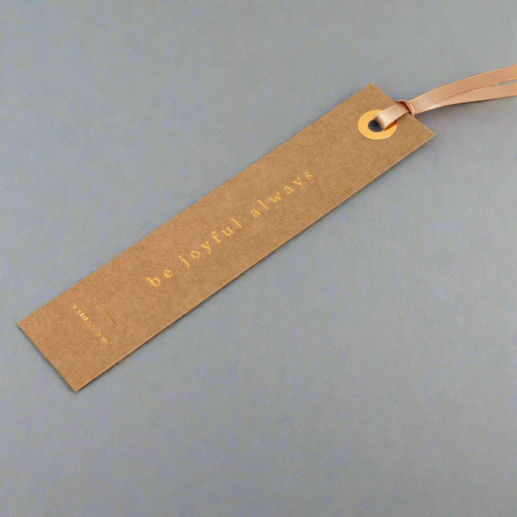 Book with a brown paper bookmark reading 'be joyful always' on a beige background
