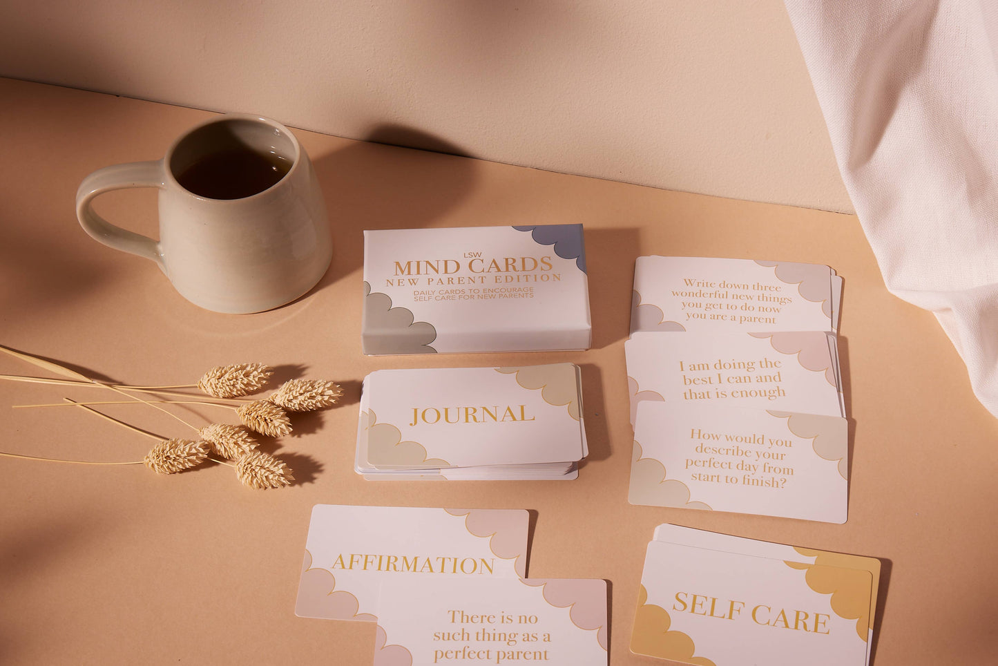 Set of cards labeled 'Mind Cards', 'Journal', 'Affirmation', and 'Self Care' on a beige surface with a mug and dried plants.