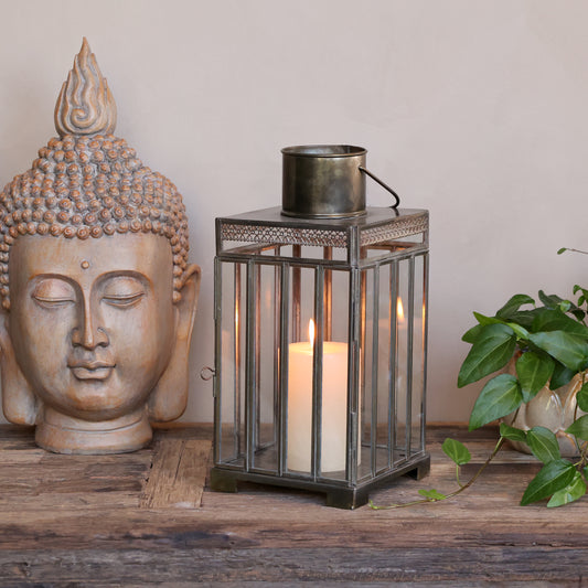Decorative lantern with candle, Buddha head, and plant on a wooden surface.