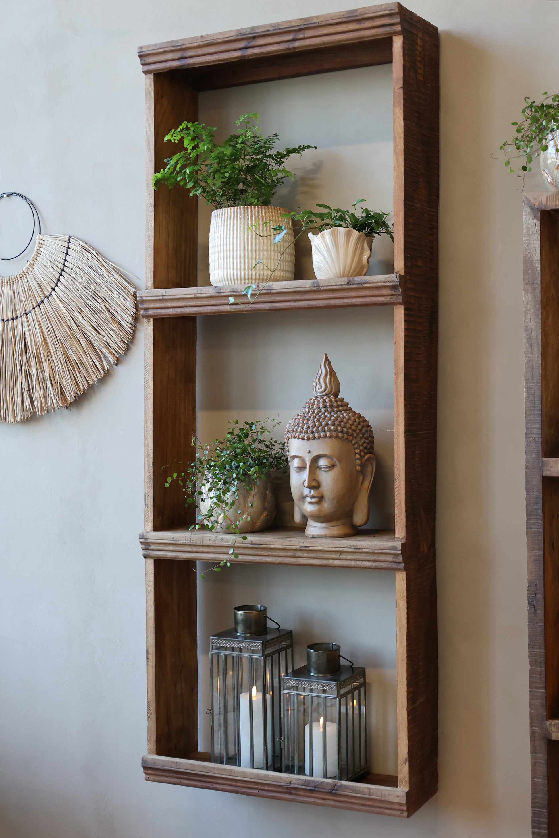 Wooden shelf with decorative items including a Buddha head and plants against a neutral wall.