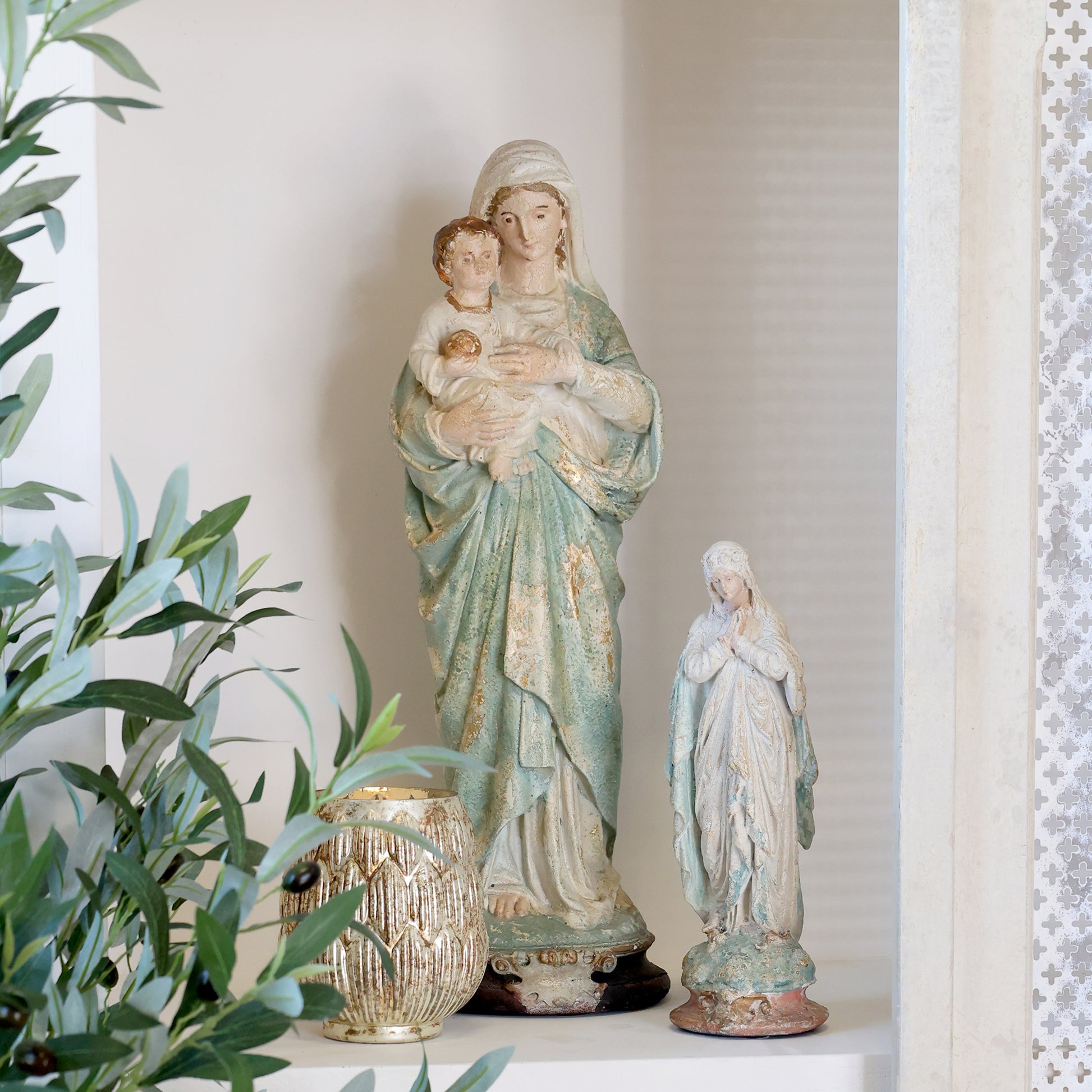 Statue of a woman holding a child with another smaller statue beside it, on a surface with plants.