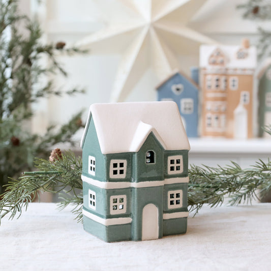 Decorative house-shaped candle with snow on top, surrounded by Christmas trees and other holiday decorations.