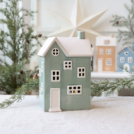 Decorative house-shaped candle holder with a snowy roof, surrounded by Christmas decorations.