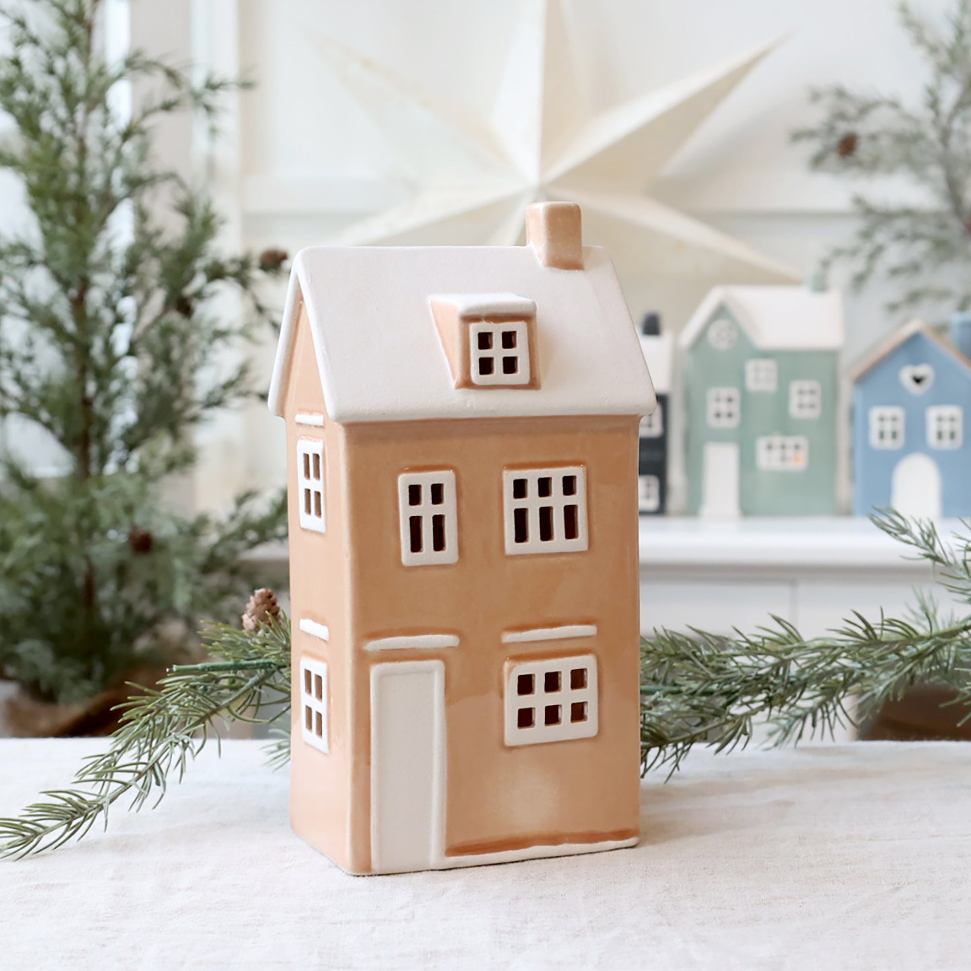 Decorative house-shaped lantern with a snowy roof, surrounded by Christmas trees and other similar houses.