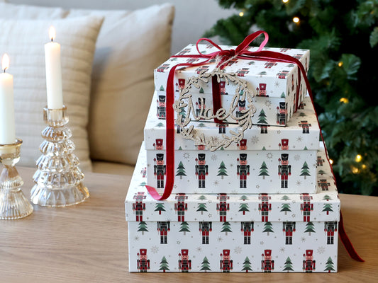 Stack of Christmas-themed gift boxes with a red ribbon on a table with candles and a tree in the background.