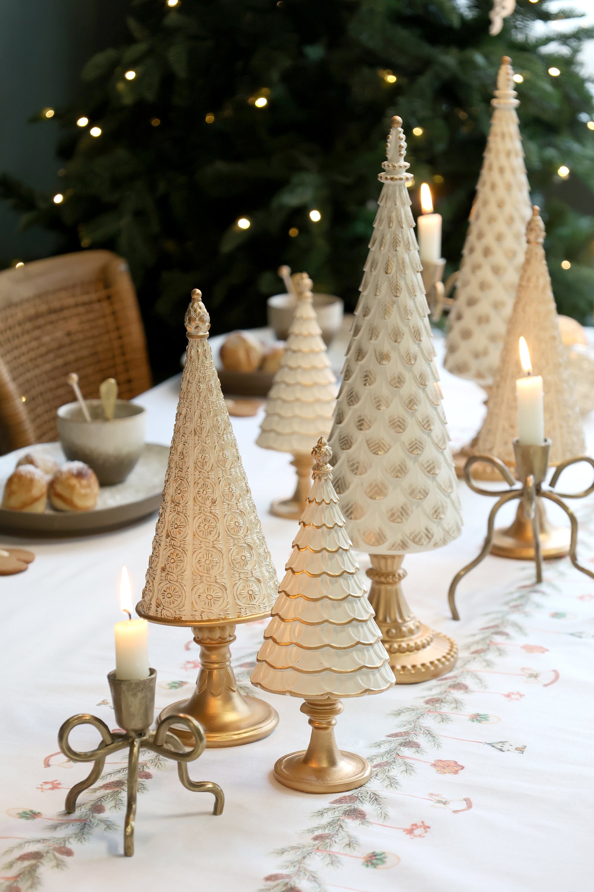 Decorative Christmas trees on a table with candles and a Christmas tree in the background.