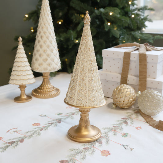 Decorative gold and white Christmas trees on a table with a Christmas tree and presents in the background.