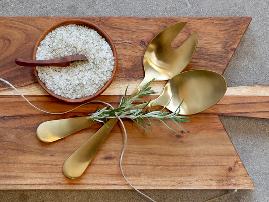 Set of gold spoons on a wooden cutting board with a small bowl of salt and a sprig of rosemary.