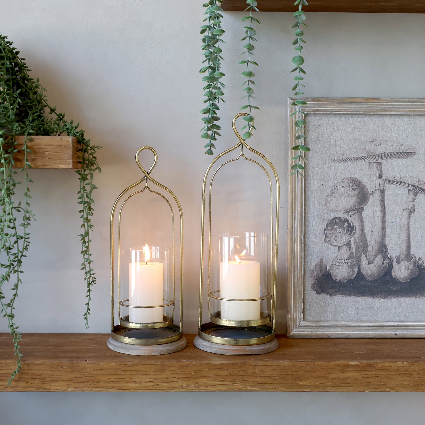 Two candle holders with candles on a wooden shelf with plants and a framed picture in the background.