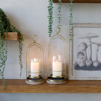 Two candle holders with candles on a wooden shelf with plants and a framed picture in the background.