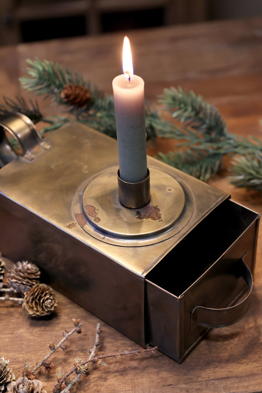 Decorative candle holder with a lit candle on a wooden surface with greenery and pine cones.