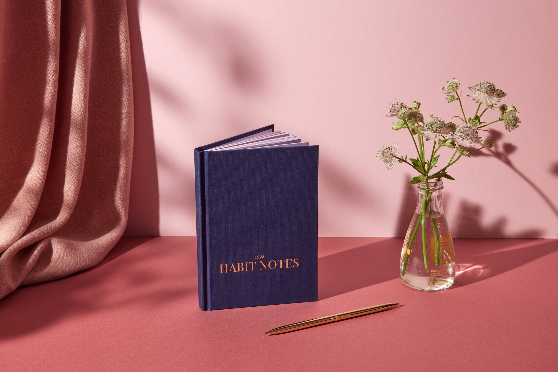 Blue notebook titled 'Habit Notes' with a pen and small vase of flowers on a pink surface.