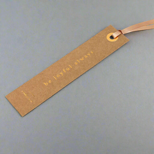 Book with a brown paper bookmark reading 'be joyful always' on a beige background