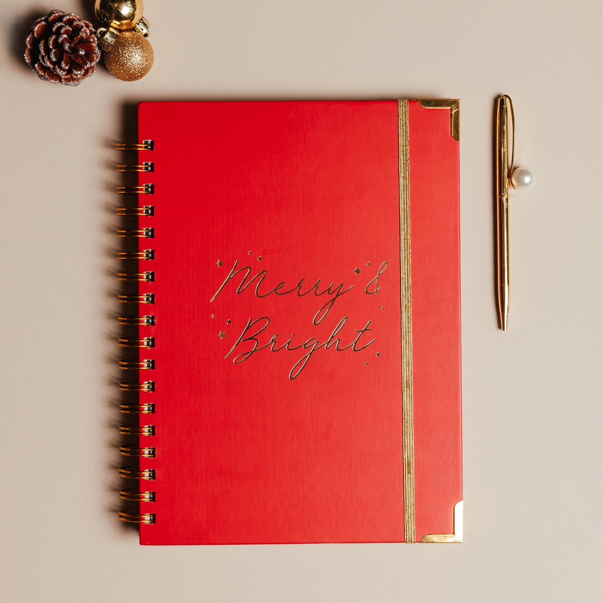 A red spiral-bound notebook with gold corner accents and the words 'Merry & Bright' in cursive on the cover, styled on a beige background with a gold pen and small holiday ornaments.