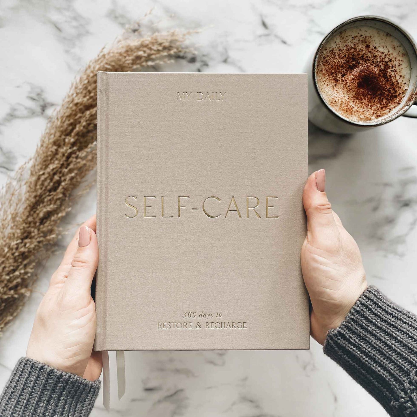 Beige 'Self-Care' journal held by hands with a cup of coffee and decorative elements on a marble surface.