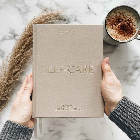 Beige 'Self-Care' journal held by hands with a cup of coffee and decorative elements on a marble surface.