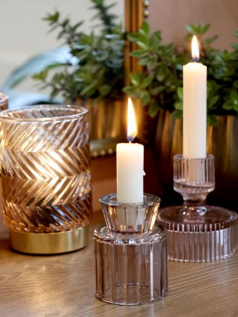 Three candle holders with lit candles on a reflective surface, with plants in the background. Generosa Home